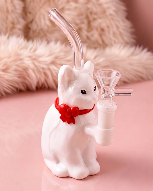 A white cat-shaped glass water pipe with a curved clear glass mouthpiece, a clear glass funnel attachment, and a red bow detail around the neck, set against a pink background with a fuzzy beige surface.