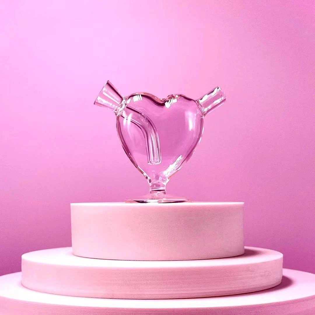 Clear heart-shaped glass blunt bubbler with curved internal downstem and small pedestal base against a pink background.