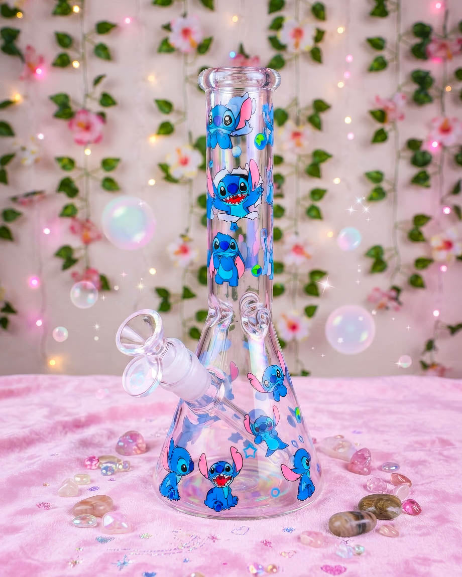 Clear 10-inch beaker bong decorated with colorful blue alien characters, hearts, and stars, featuring a 14mm glass bowl on a pink floral background.