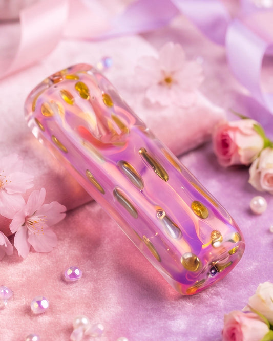 Iridescent pink and lavender glass pipe with gold gem-like sprinkle accents displayed on satin among flowers, pearls, and crystals.